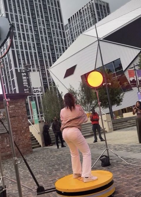 Melissa Steel performing on the yellow On Da Booth platform outdoors between city buildings, with lights and camera stands around her
